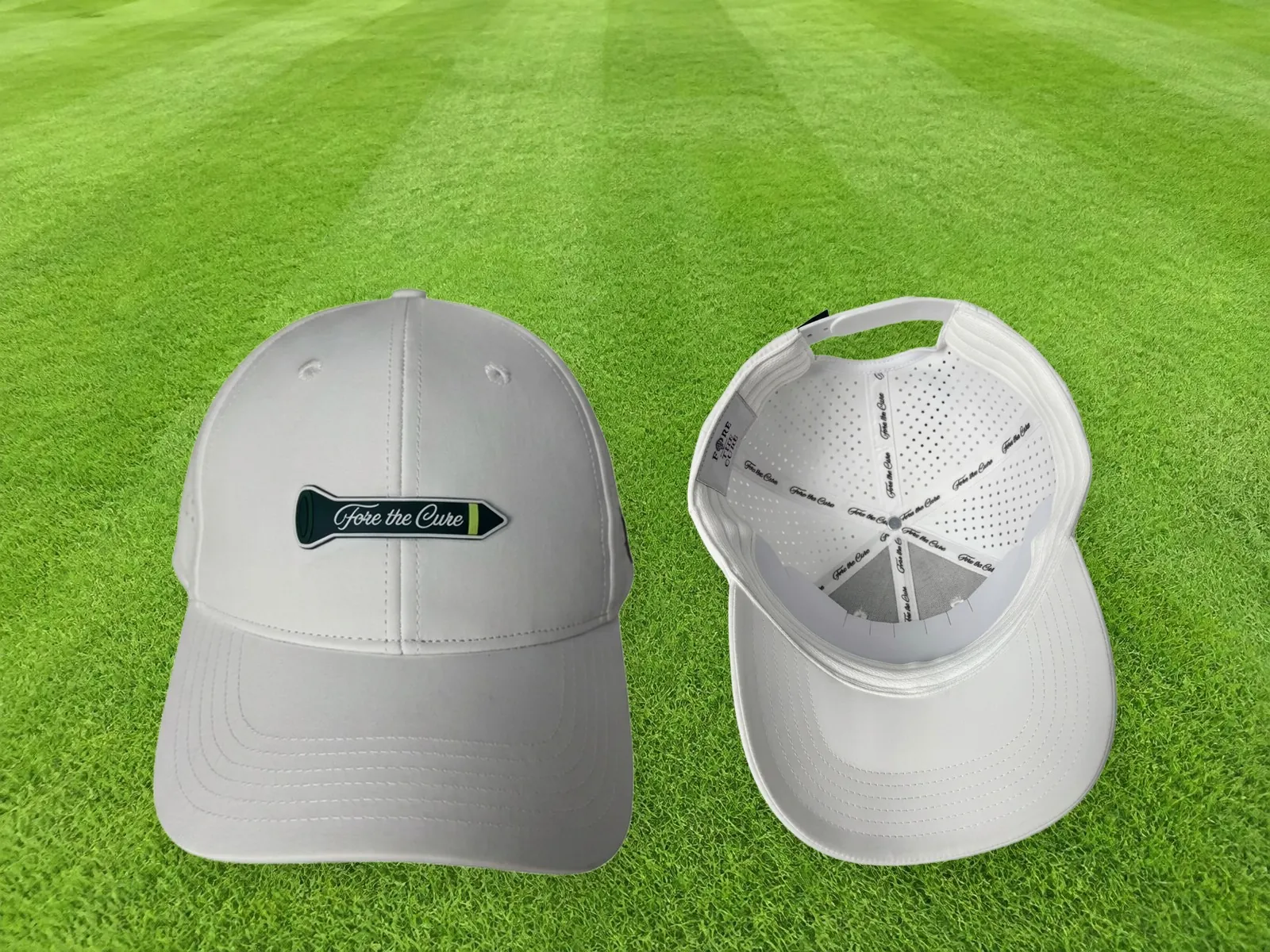 Two white Fore the Cure fundraiser caps sitting side by side on golf course grass